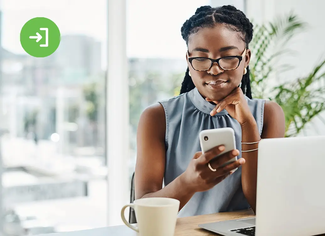 A woman wearing glasses sits at a desk with a laptop open in front of her, smiling while holding and looking at a smartphone.