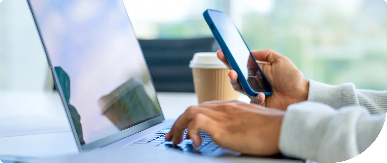 Close-up of a person’s hands using a laptop while holding a smartphone, with a coffee cup on the desk in the background.