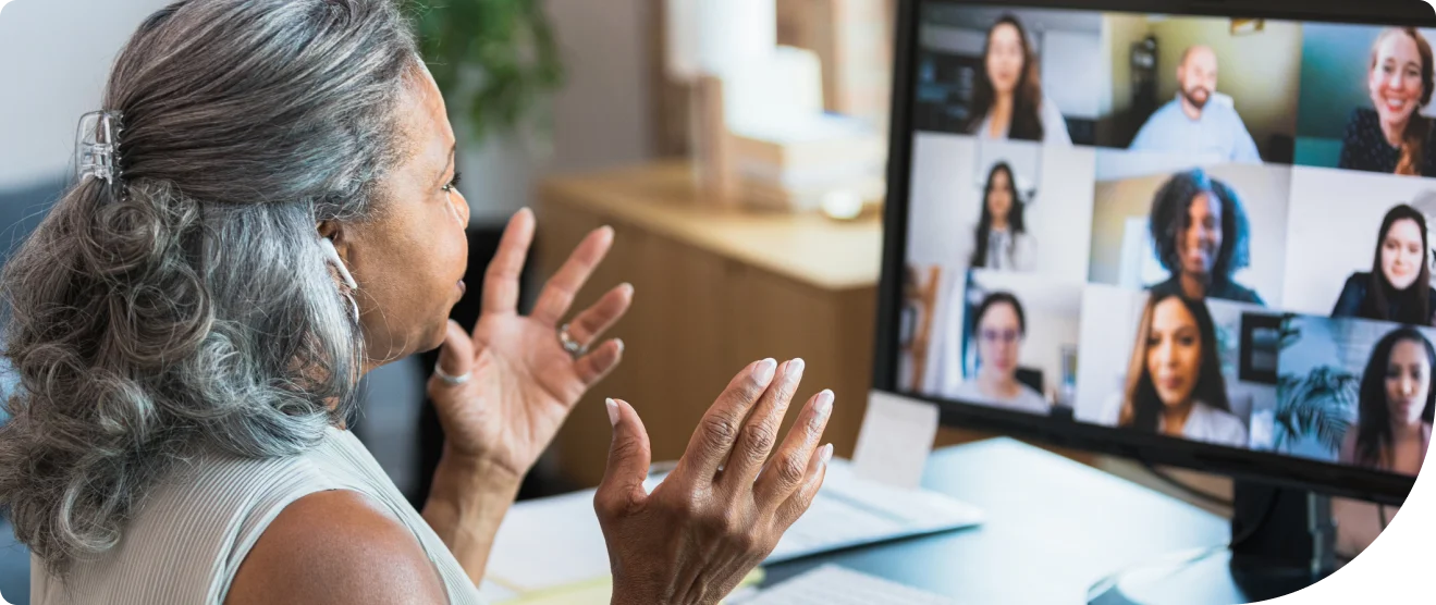 Woman with gray hair on a video call, gesturing while talking to multiple colleagues displayed in a grid on a computer monitor