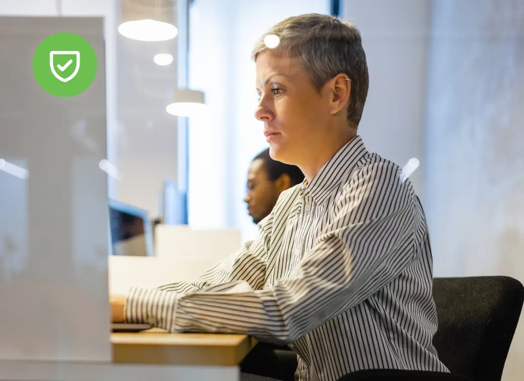 Woman with short gray hair working at a computer in a bright office environment with modern lighting and other employees nearby