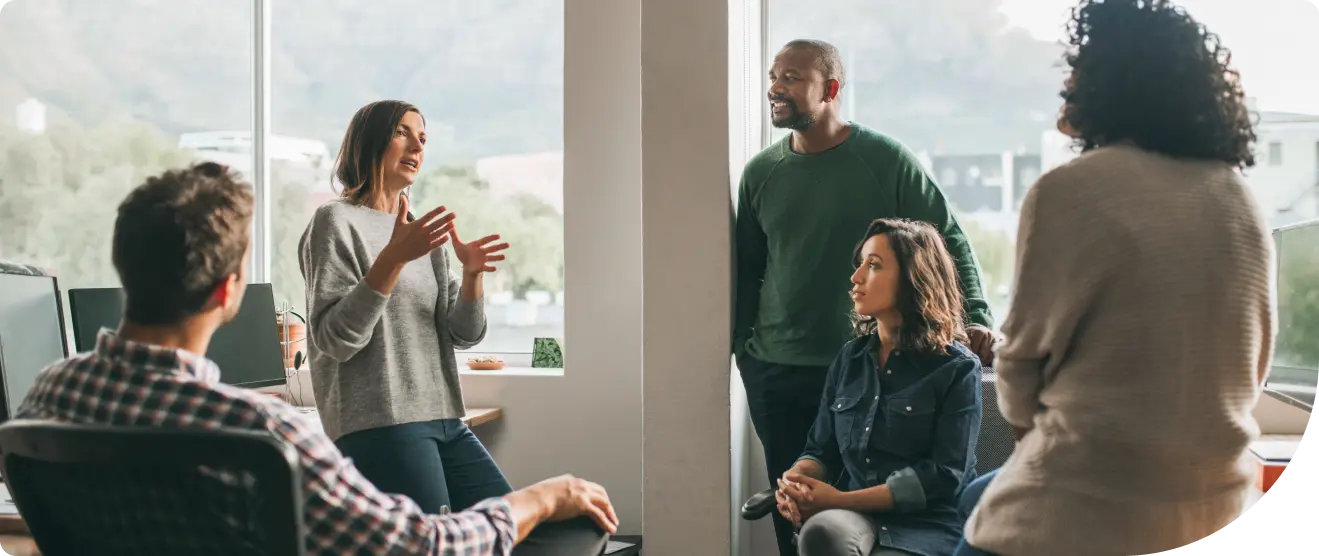 A group of colleagues gathered by office windows, with one woman standing and speaking while the others listen and engage in discussion.