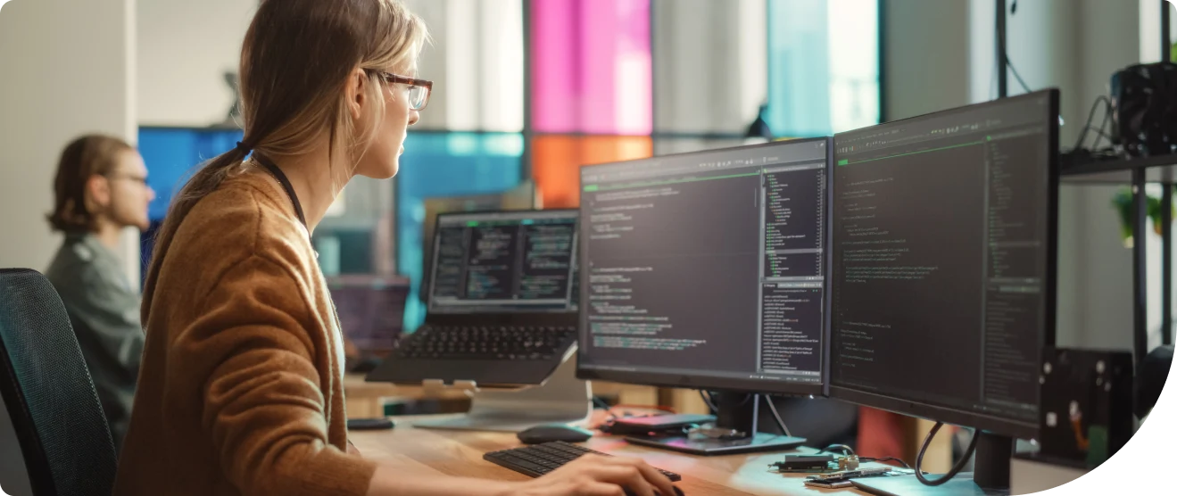 A woman works at a desk with multiple monitors displaying code in a modern office