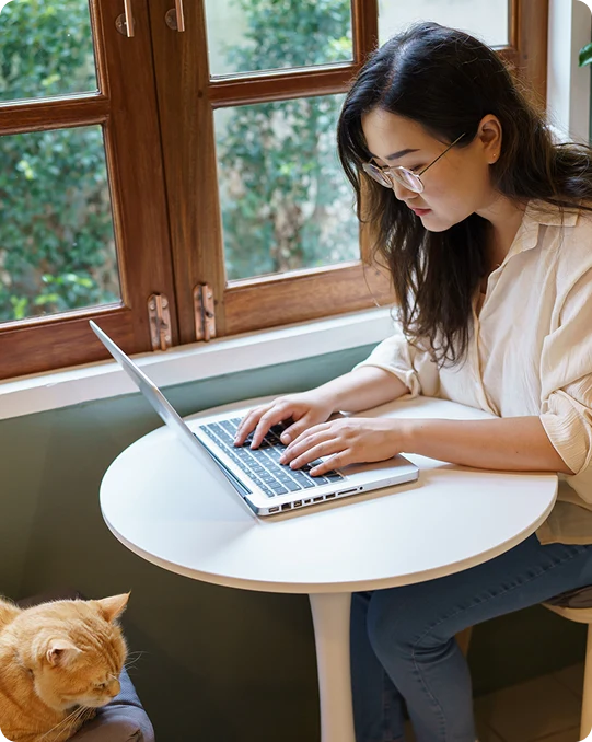 Woman types on a laptop at a small table by a window with a cat sitting nearby
