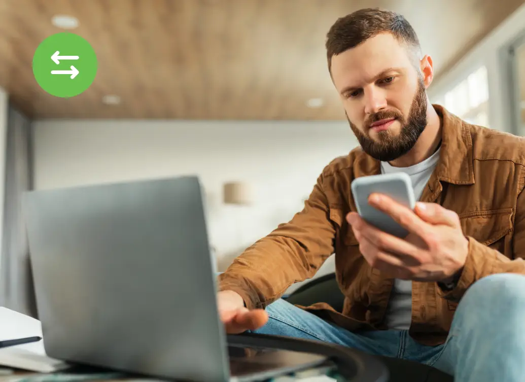Man sitting at a table using a laptop while holding a smartphone, appearing focused as he works in a home or office setting