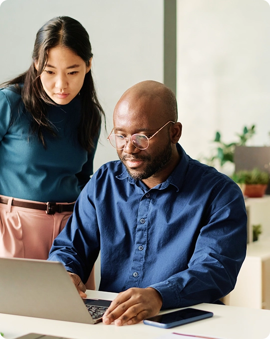 Two coworkers focus on a laptop screen in a modern office, one seated and typing while the other observes the screen