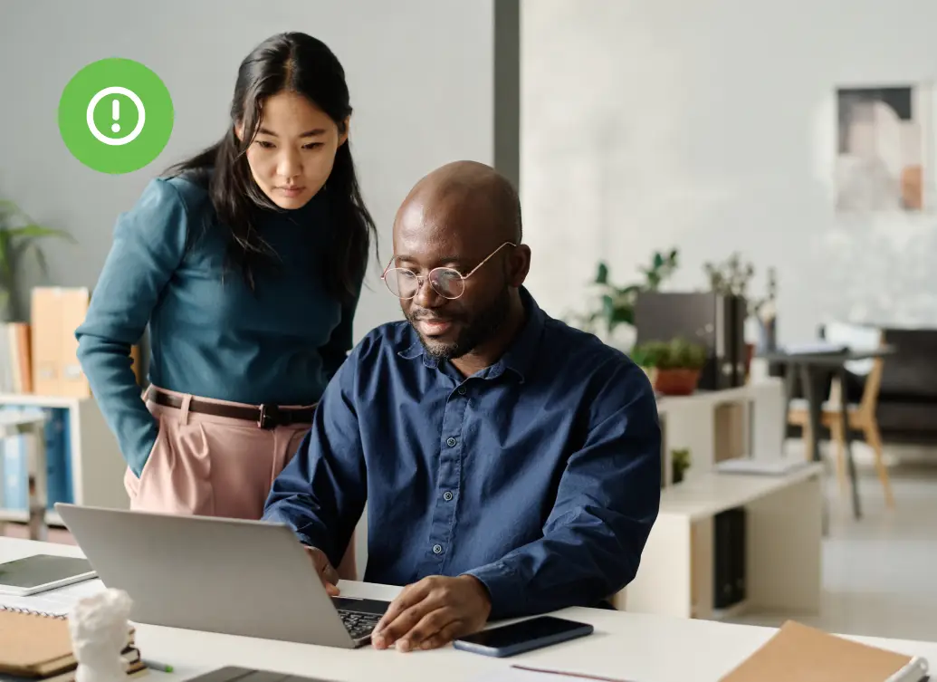 Two coworkers focus on a laptop screen in a modern office, one seated and typing while the other observes the screen