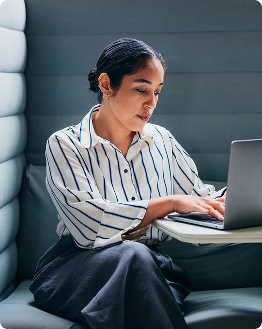 Woman in a striped shirt types on a laptop while seated in a modern booth with soft gray walls