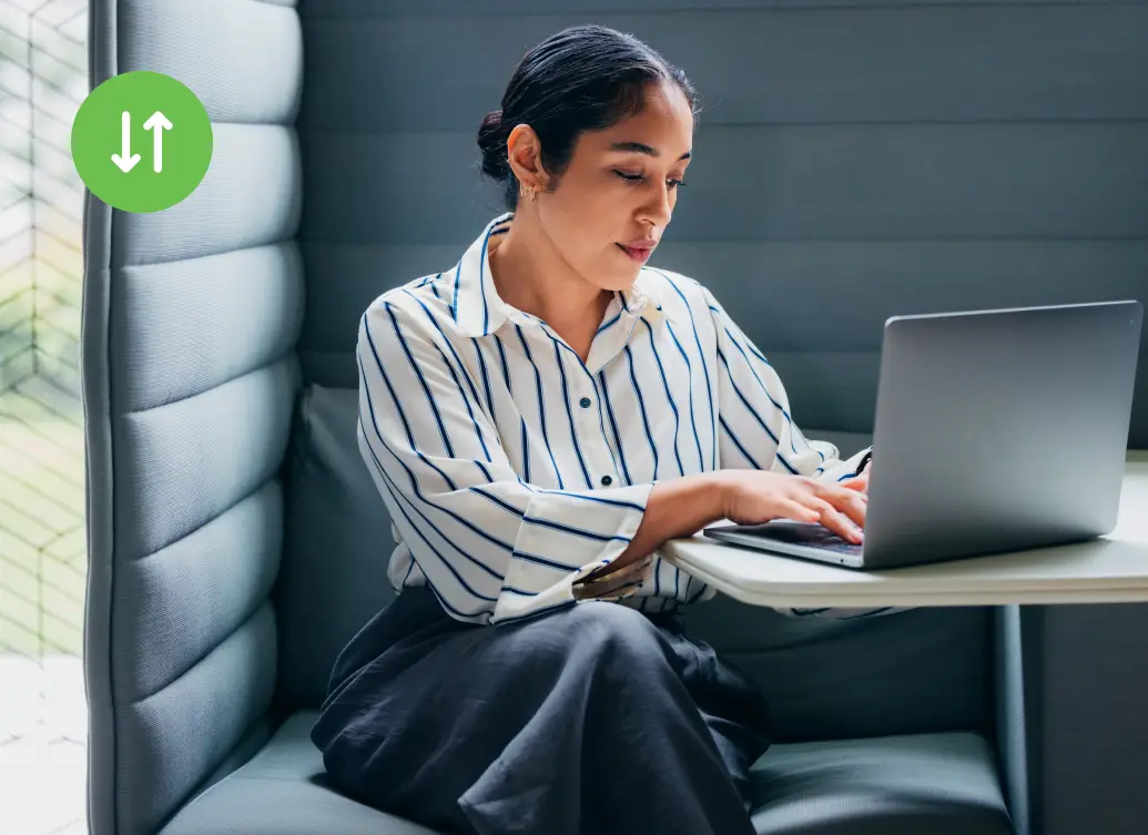 Woman in a striped shirt types on a laptop while seated in a modern booth with soft gray walls