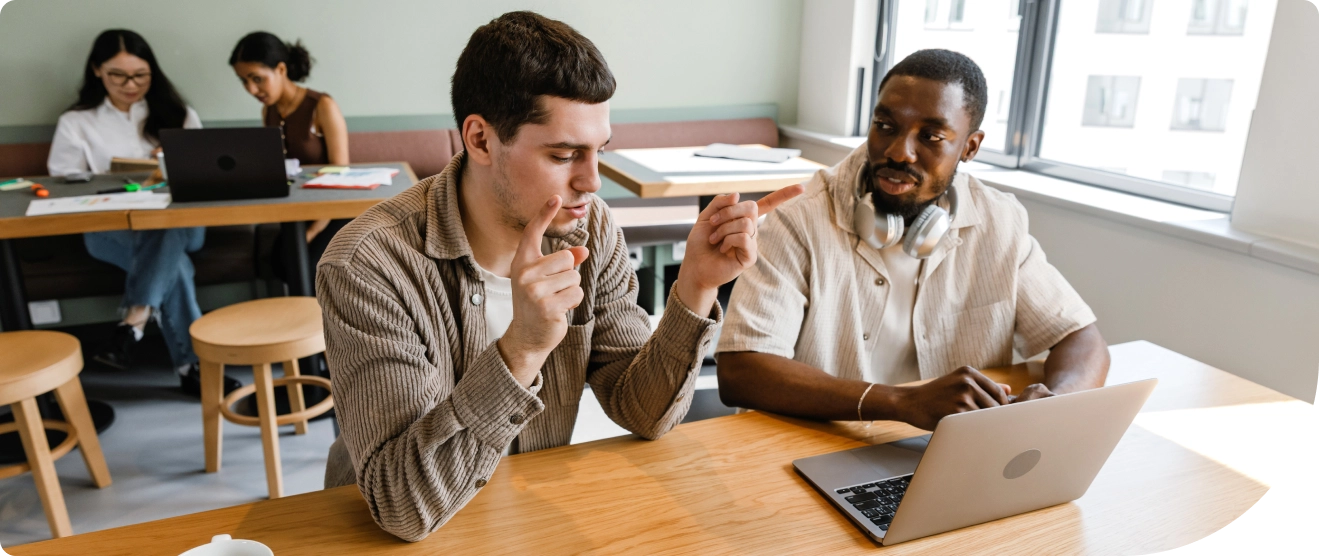 Two colleagues collaborate at a laptop in a bright office while two others work together in the background