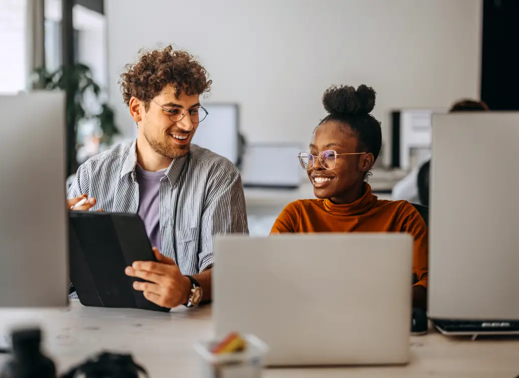 Two coworkers smiling and talking at their desks, one holding a tablet and the other typing on a laptop in a modern office setting.