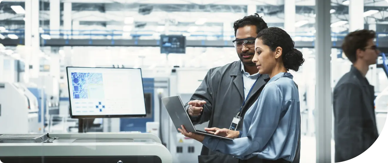 Colleagues in a factory discuss data on a laptop near computer screens and production equipment