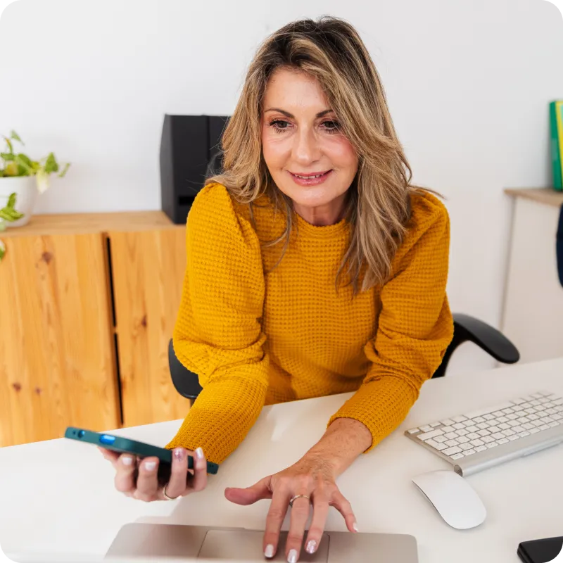 Person in yellow sweater using a laptop and holding a phone at a desk